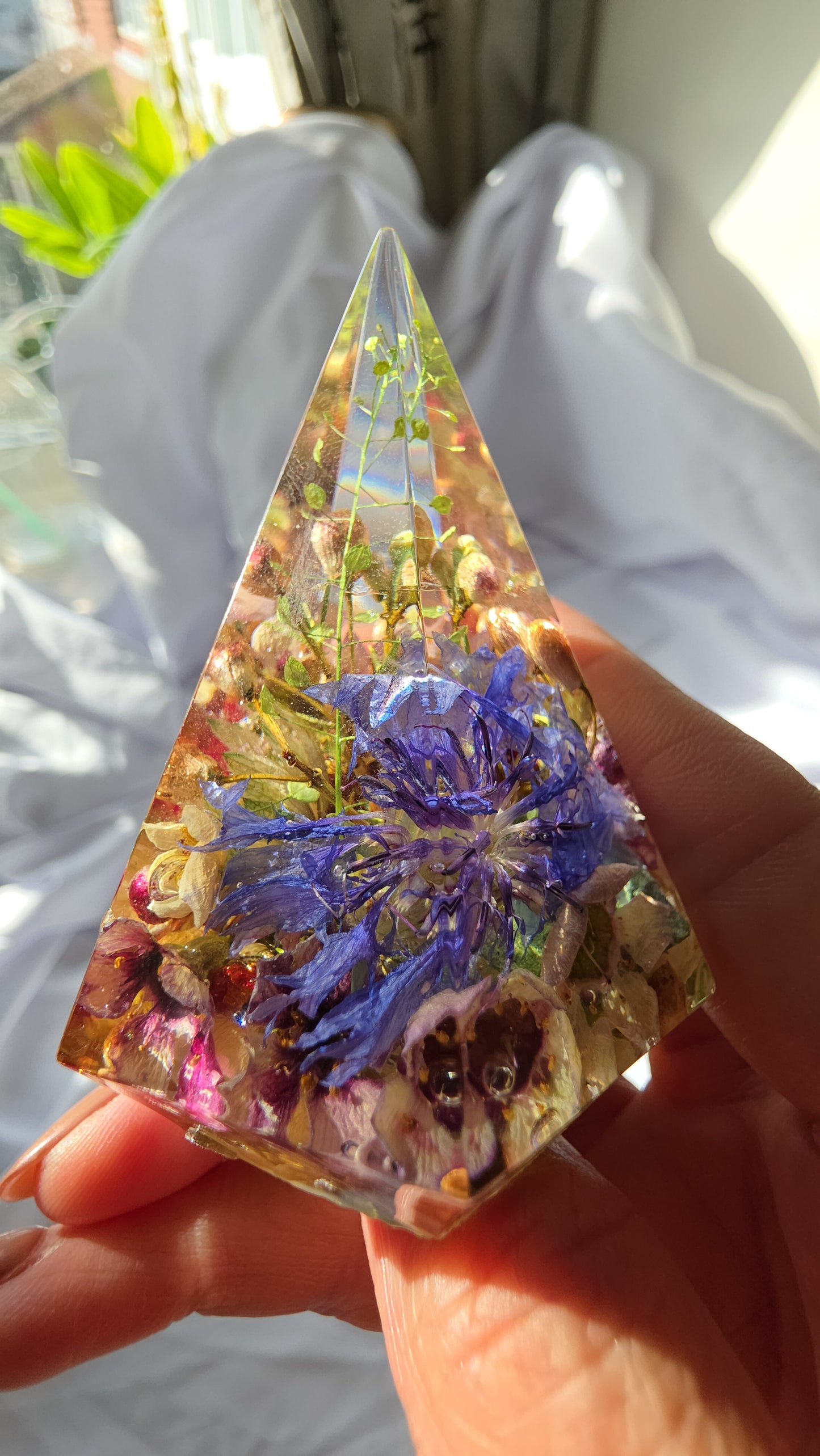 Close-up of a small resin ring holder containing preserved flowers, shown held in hand, with layered petals and greenery visible inside the clear pyramid-shaped piece.