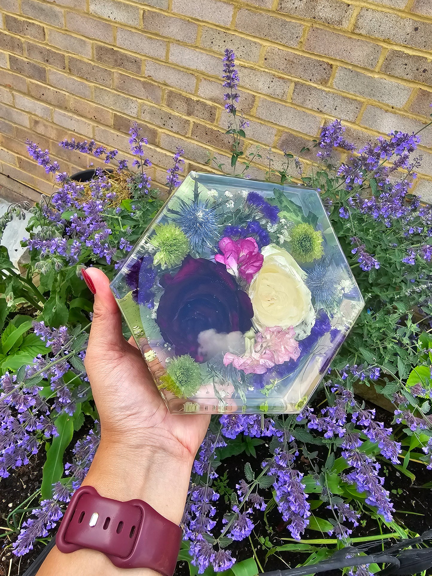 A hand holding a custom resin hexagon block with a preservation of flowers inside, set against a background of purple lavender.