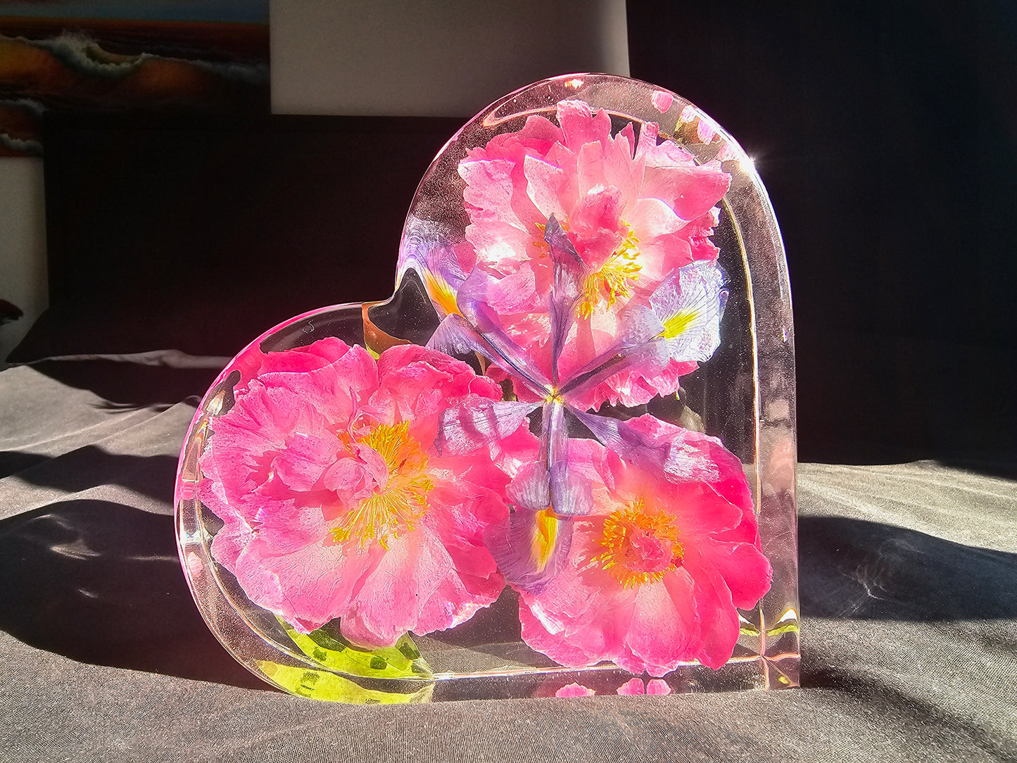 A heart-shaped resin block with preserved pink flowers and foliage, placed on a flat surface with a source of light illuminating it.