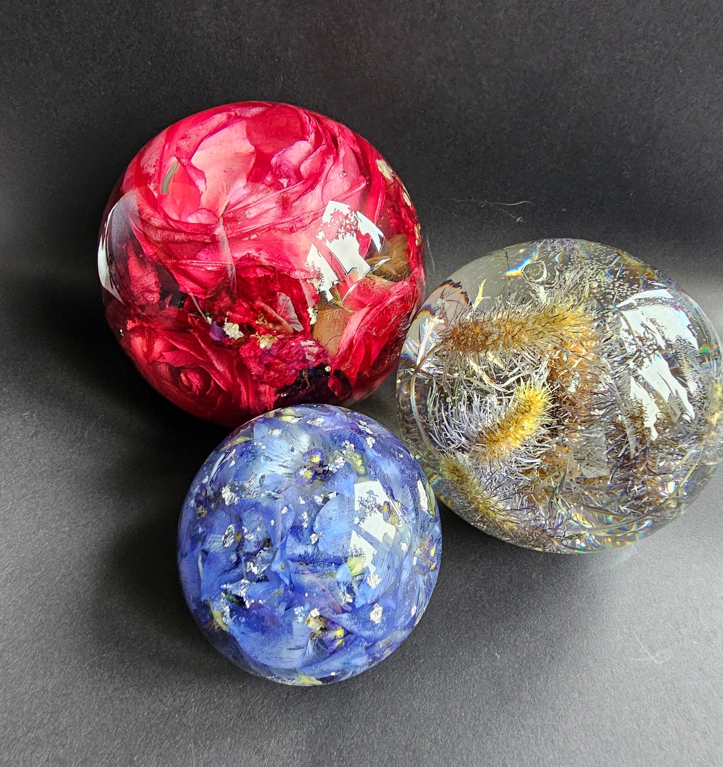 Three handcrafted resin flower preservation spheres in different sizes, with each containing different flowers. The spheres are transparent, showcasing the colorful flowers inside.